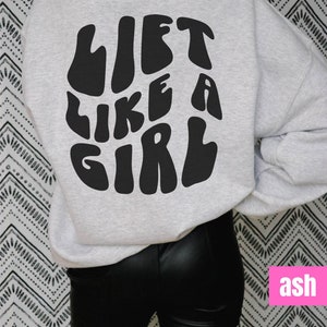 Puede incluir: Una sudadera gris con el texto "Lift Like A Girl" impreso en negro en la espalda. El texto est&aacute; en una fuente retro y ondulada. La sudadera la lleva puesta una persona que lleva pantalones negros.