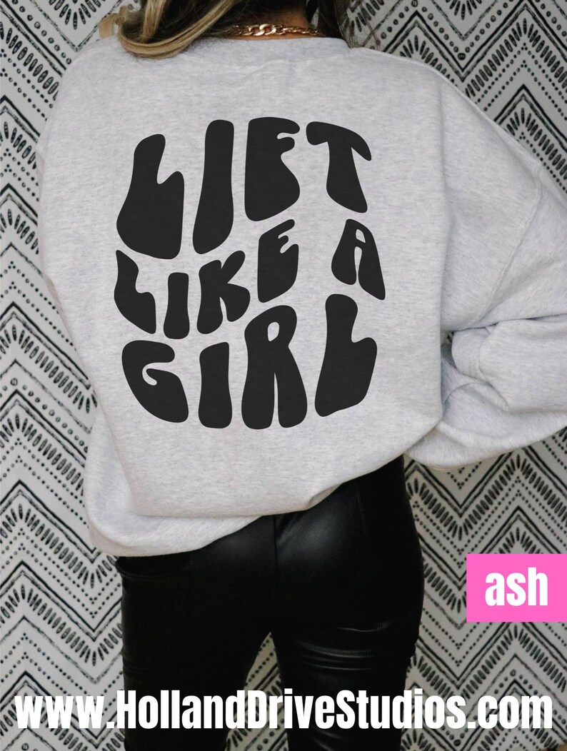 Puede incluir: Una sudadera gris con el texto "Lift Like A Girl" impreso en negro en la espalda. El texto est&aacute; en una fuente retro y ondulada. La sudadera la lleva puesta una persona que lleva pantalones negros.