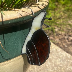 May include: A stained glass snail ornament featuring a translucent white body and a dark brown, textured shell with black lead lines. The snail is attached to the rim of a green and beige ceramic planter containing green grass blades.