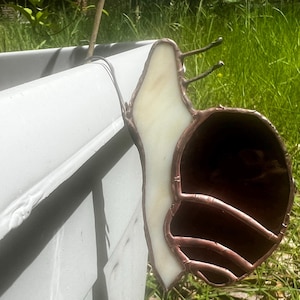 May include: A stained glass snail ornament featuring a cream-colored body and a dark brown, spiraled shell constructed with soldered metal lines. The snail is positioned on the edge of a light gray plastic planter, showcasing its curved form.