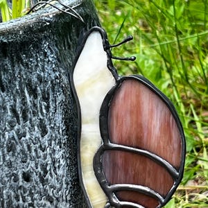 May include: A stained glass snail ornament featuring a white, opalescent body and a reddish-brown, swirled shell. The glass pieces are joined by dark metal solder lines, and the snail has two thin, dark metal antennae extending from its head.