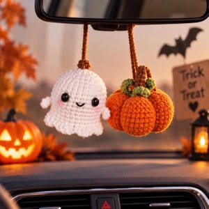 Könnte beinhalten: Ein Paar hängende Häkelornamente für den Rückspiegel im Auto, bestehend aus einem weißen Geist mit schwarzen Augen und rosa Wangen sowie einem orangefarbenen Kürbis mit braunem Stiel und grünen Blättern, befestigt an braunen Garnschnüren.