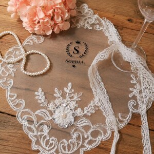 May include: A clear acrylic sign featuring the name Sophia in black text with a crown icon, decorated with white lace trim, a pearl flower accent, and a string of white pearls. A glass stemware base sits on the right side of the sign.