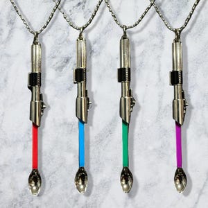 May include: Four silver-toned metal pendants on ball chains, each shaped like a lightsaber hilt with a small spoon attached at the bottom. The spoons feature colored stems in red, blue, green, and purple, with metallic silver bowls at the ends.