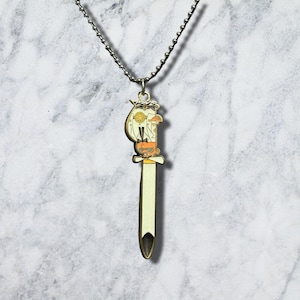 May include: A metal pendant necklace featuring a long, sword-shaped charm with a cream-coloured blade and a decorative hilt. The hilt displays a stylised orange and white character design with a circular emblem, suspended from a silver ball chain.