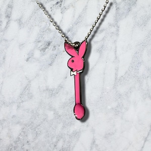 May include: A silver-toned metal ball chain necklace featuring a pendant shaped like a long, narrow spoon. The handle and bowl of the spoon are filled with bright pink enamel, and the top features a pink rabbit head silhouette with a white bowtie.