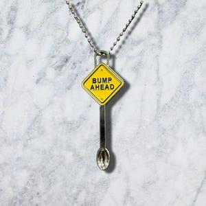 Może przedstawiać: Srebrzysty naszyjnik z metalu z zawieszką w kształcie znaku drogowego z żółtym tłem i czarnym napisem BUMP AHEAD. Zawieszka jest przymocowana do srebrnego łańcuszka kulkowego i posiada małe przedłużenie w kształcie łyżeczki u podstawy.