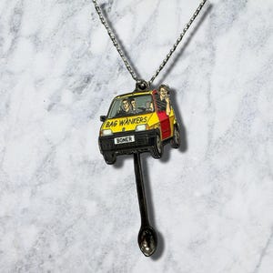 May include: A silver-toned metal necklace featuring a pendant shaped like a yellow and red car with the text BAG WANKERS on the hood and BONER on the license plate. The car pendant is attached to a small spoon-shaped charm hanging below it.