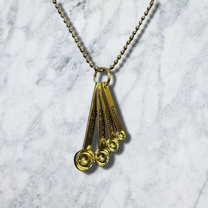 Puede incluir: Un collar de metal dorado con un colgante compuesto por cuatro cucharas medidoras en miniatura unidas a una cadena de bolas. Cada cuchara está grabada con texto, incluyendo las palabras bump, me y lines, colgadas de una argolla circular.
