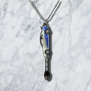 May include: A silver-toned metal necklace featuring a pendant shaped like a sports car with blue racing stripes on the side. The car is attached to a small spoon handle engraved with the text FORGET ABOUT IT CUH in white capital letters.
