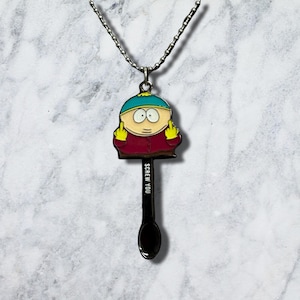 May include: A silver-toned ball chain necklace featuring a pendant shaped like a spoon. The handle of the spoon is black with the white text "SCREW YOU." The bowl of the spoon features a colourful enamel character with a teal hat and red jacket.