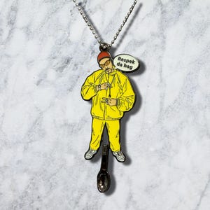 May include: A metal pendant necklace featuring a cartoon figure wearing a yellow tracksuit, a red cap, and glasses. The figure is making a hand gesture and has a speech bubble that reads Respek da bag. A small metal spoon hangs from the figure's feet.