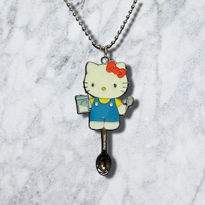 Może przedstawiać: Naszyjnik z zawieszką z metalu w kolorze srebrnym przedstawiający postać Hello Kitty w niebieskich ogrodniczkach i żółtej koszulce, trzymającą mały kubek i łyżkę. Postać jest przymocowana do podstawy w kształcie łyżki z wygrawerowanym napisem Hello Kitty.
