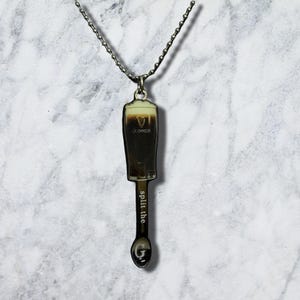 May include: A silver-toned metal pendant necklace featuring a miniature Guinness pint glass design with a creamy white head and dark body. The stem of the pendant is shaped like a spoon with the text "split the" and a "G" printed on the bowl.