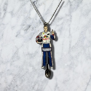 May include: A silver-toned metal necklace featuring a pendant shaped like a race car driver in a white and blue suit with the text "Wonder" and "WAKE UP AND SNIFF EXCELLENCE" printed on the front, attached to a delicate silver ball chain.