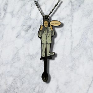 May include: A metal pendant necklace featuring a character in a light grey suit giving two thumbs up. A speech bubble reads "nice bump!" and a small sign reads "GREAT SUCCESS". The pendant is attached to a dark metal ball chain necklace.