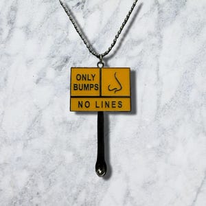Op de afbeelding: Een zilverkleurige metalen ketting met een rechthoekige gele hanger met zwarte tekst die ONLY BUMPS en NO LINES leest, vergezeld van een afbeelding van een neus. Een kleine zilverkleurige lepel is aan de onderkant van de hanger bevestigd.