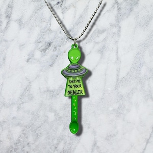 Può includere: Una collana con ciondolo in metallo verde raffigurante una testa di alieno sopra un disco volante e un piccolo manico di cucchiaio. Il ciondolo riporta la scritta "TAKE ME TO YOUR DEALER" in nero. L'oggetto è appeso a una catenina a palline argentata.