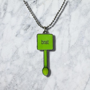May include: A silver-toned ball chain necklace featuring a pendant shaped like a square sign with a vertical stem and a small circular base. The square portion is bright lime green with the word brat printed in black lowercase letters.