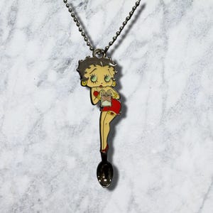 May include: A silver-toned metal pendant necklace featuring a cartoon character with black hair, large eyes, and gold hoop earrings, wearing a red dress and holding a small bag. The pendant is shaped like a spoon at the base and hangs on a ball chain.