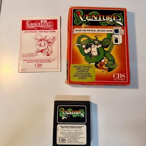 May include: A Venture video game set for the Atari Video Computer System, including a rectangular cardboard box with red and orange graphics, a black plastic game cartridge, and a white paper instruction manual with red text.
