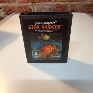 May include: A black Atari 2600 video game cartridge for Star Raiders. The label features the game title in orange text, instructions to use with a joystick controller and video touch pad, the Atari logo, and a space combat illustration.