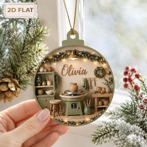 May include: A flat, circular, sage green Christmas ornament featuring a detailed miniature pottery studio scene. The interior displays a potter's wheel, shelves with ceramic vessels, a stool, and the name Olivia in script. It hangs by a gold cord.
