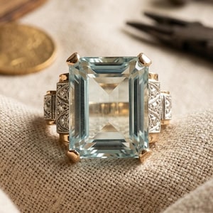 May include: A gold ring featuring a large, emerald-cut, light blue gemstone set in the center. The band is accented with small, clear, round-cut stones embedded in a decorative metal setting on both sides of the central light blue gemstone.