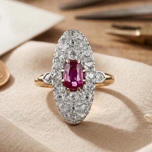 Peut inclure: Une bague de forme ovale avec une pierre précieuse rose foncé au centre, entourée de nombreuses petites pierres claires et scintillantes serties dans une monture métallique. La bague possède un anneau fin en or sur un tissu clair.