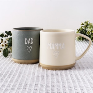 Könnte beinhalten: Zwei Keramikbecher mit matter Oberfläche. Der linke Becher ist dunkelgrau mit dem Wort DAD und einem Herzumriss in Weiß. Der rechte Becher ist cremefarben mit dem Wort MAMMA und einem Herzumriss in Weiß. Beide haben einen beigen Boden.