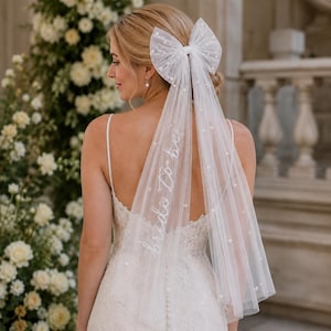 May include: A white tulle bridal veil with scattered pearls and the text "bride to be" in script. The veil is attached to a large bow. The bride is wearing a white spaghetti strap dress.