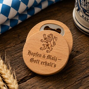 Könnte beinhalten: Holz-Flaschenöffner und Untersetzer-Set. Der Öffner ist rund mit einem silbernen Metallöffner. Der Untersetzer hat ein Löwenwappen und den Text "Hopfen & Malz - Gott erhalt's" eingraviert.