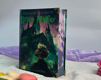 May include: A hardcover book titled The Disappearances of Draco Malfoy by Speechwriter, featuring a dark illustration of a castle and a person in the foreground. The book has metallic gold corner protectors on the top and bottom edges of the cover.