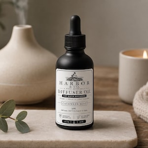 Op de afbeelding: Een zwarte fles diffuserolie met een druppelaar, met het label "Harbor & Co. Diffuser Oil - Cashmere Mist". De fles staat op een marmeren dienblad, naast een brandende kaars in een beige houder, een beige diffuser en takjes eucalyptus.