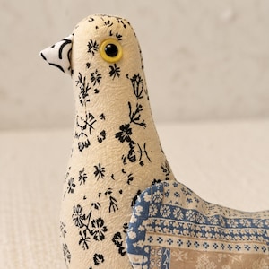 May include: A stuffed fabric bird toy featuring a cream-colored body with a black floral print, a white beak with black markings, and a yellow eye. The wing is made of a patchwork fabric with blue and tan geometric and floral patterns.
