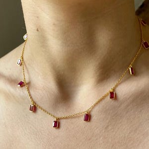 May include: A gold chain necklace featuring multiple small, rectangular, deep red gemstones suspended at intervals along the delicate link chain. The stones are set in gold bezels, creating a consistent pattern of red accents around the neck.