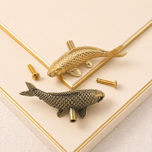May include: Two koi fish-shaped cabinet knobs in metallic finishes, one bright gold and one antique bronze. Each knob features detailed scale textures, fins, and a cylindrical mounting post. Two small gold-coloured metal screws are included.