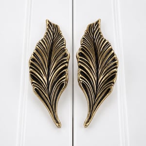 Peut inclure: Deux poignées de placard assorties en forme de feuilles allongées. Chaque poignée présente une finition en laiton antique métallique avec des détails sombres en creux qui soulignent les motifs de nervures en relief le long de la feuille.