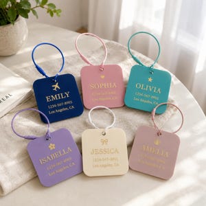 Puede incluir: Seis etiquetas de equipaje cuadradas en azul marino, rosa, verde azulado, lavanda, crema y malva, cada una con un lazo de alambre dorado. Cada etiqueta muestra un nombre, un número de teléfono, una ciudad y un pequeño icono decorativo.