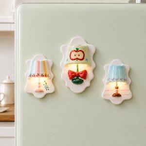 Może przedstawiać: Trzy ceramiczne magnesy w kształcie kinkietów z podświetlanymi kloszami. Lewa lampa ma pastelowe paski i motywy wiśni. Środkowa lampa ma wzór jabłka i czerwoną kokardę. Prawa lampa ma niebieski, falowany klosz z różowymi liniami.