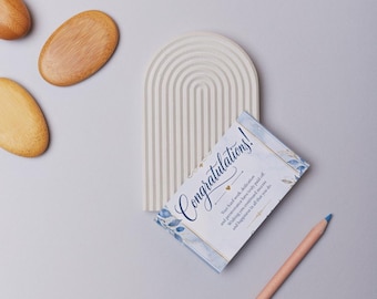 May include: A beige arched tray with a ribbed texture, three smooth oval wooden stones, a rectangular card with blue floral accents and the text Congratulations! Your hard work, dedication and perseverance have truly paid off.