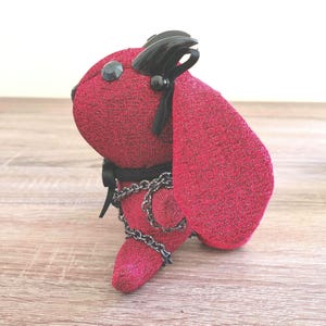 May include: A plush rabbit toy crafted from shimmering red fabric with a black metallic sheen. The figure features black horn-like protrusions on its head, a black ribbon collar, and a decorative silver-toned chain wrapped around its body.