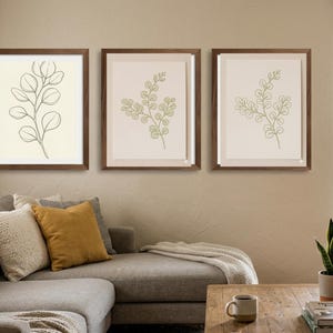Puede incluir: Tres grabados botánicos enmarcados con marcos de madera marrón. Los grabados presentan dibujos lineales de hojas de eucalipto en tonos verdes y crema. La obra de arte se exhibe sobre un sofá seccional gris con cojines decorativos.
