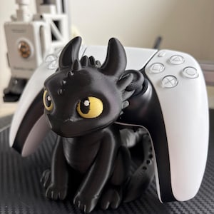 Può includere: Una statuetta di drago nera stampata in 3D con grandi occhi gialli e orecchie a punta, posizionata davanti a un controller per videogiochi bianco e nero. Il drago ha una finitura opaca liscia e siede con la coda avvolta attorno al controller.