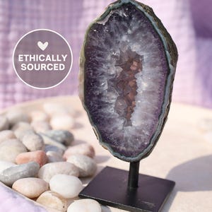 Könnte beinhalten: Eine vertikale Amethyst-Geodenscheibe mit tiefviolettem kristallinem Inneren und dunklem Außenrand, montiert auf einem schwarzen Metallständer. Eine kreisförmige weiße Grafik links enthält ein Herzsymbol und den Text ETHICALLY SOURCED.