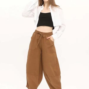 May include: A white long-sleeved button-down shirt worn open over a black cropped tank top, paired with brown wide-leg trousers featuring an elastic drawstring waistband, side pockets, and white sneakers, complemented by dark round sunglasses.