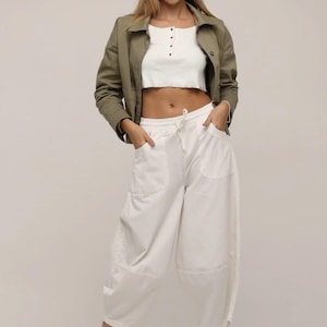 May include: An olive green collared jacket worn over a white cropped henley top and loose-fitting white drawstring trousers with side pockets. The outfit is completed with white sneakers and light-tinted glasses, creating a layered look.