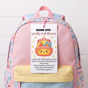 May include: A pink and light blue floral backpack with a rectangular white tag attached by a string. The tag features a cartoon backpack illustration, yellow stars, and the text: Packed with snacks and dreams, Tucked in today, right alongside your books.