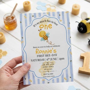 May include: A rectangular invitation card with a blue and white striped border featuring a cartoon bee, honeycomb graphics, and text that reads: It's so much fun to bee one, Ronnie's FIRST BEE-DAY, SATURDAY 6th JUNE 2pm, 221B BEE LANE, HONEY TOWN.