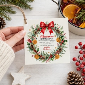 May include: A square white gift tag featuring a watercolor Christmas wreath with pine branches, red berries, pinecones, orange slices, and a red bow. The text reads: Christmas Simmer Pot, Add ingredients to a saucepan, cover with 2 cups of water, simmer on low, top up water as needed. With Love The Greens.